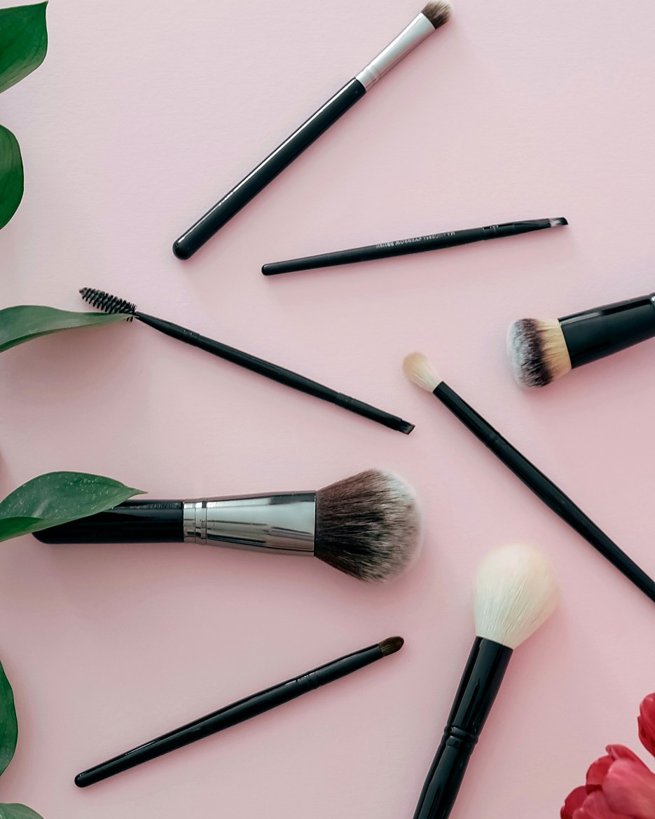 Bridal makeup brushes laid out on soft pink with green leaves